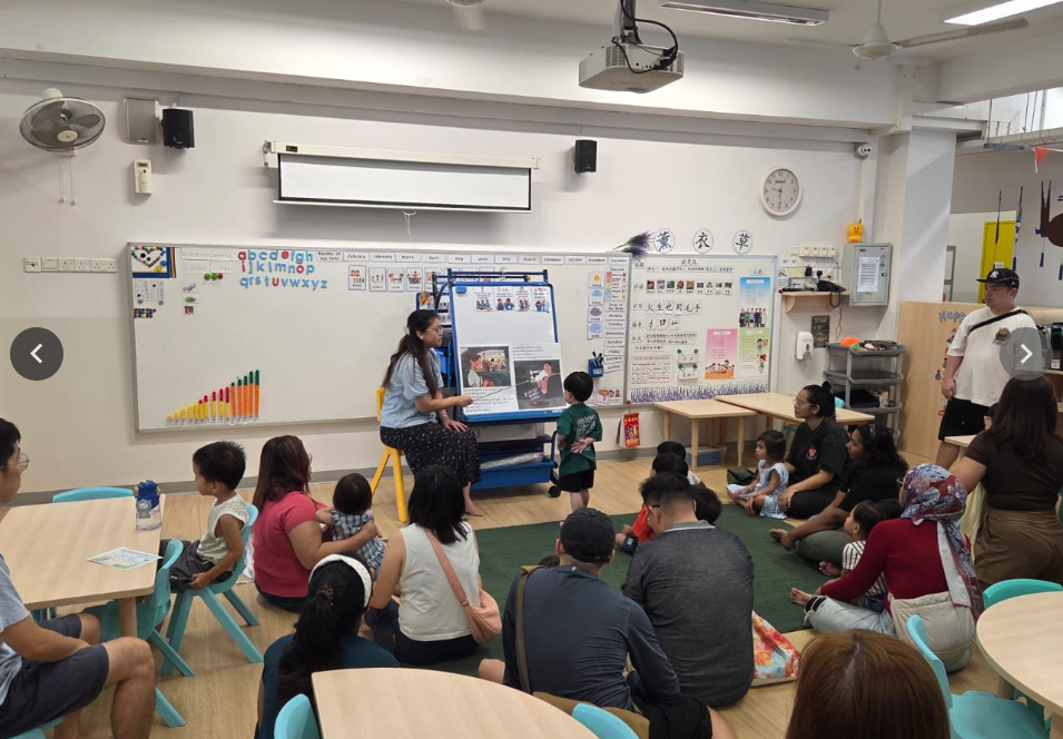 Teacher sits by a board, speaking to a child as adults and kids sit on the floor in a classroom.